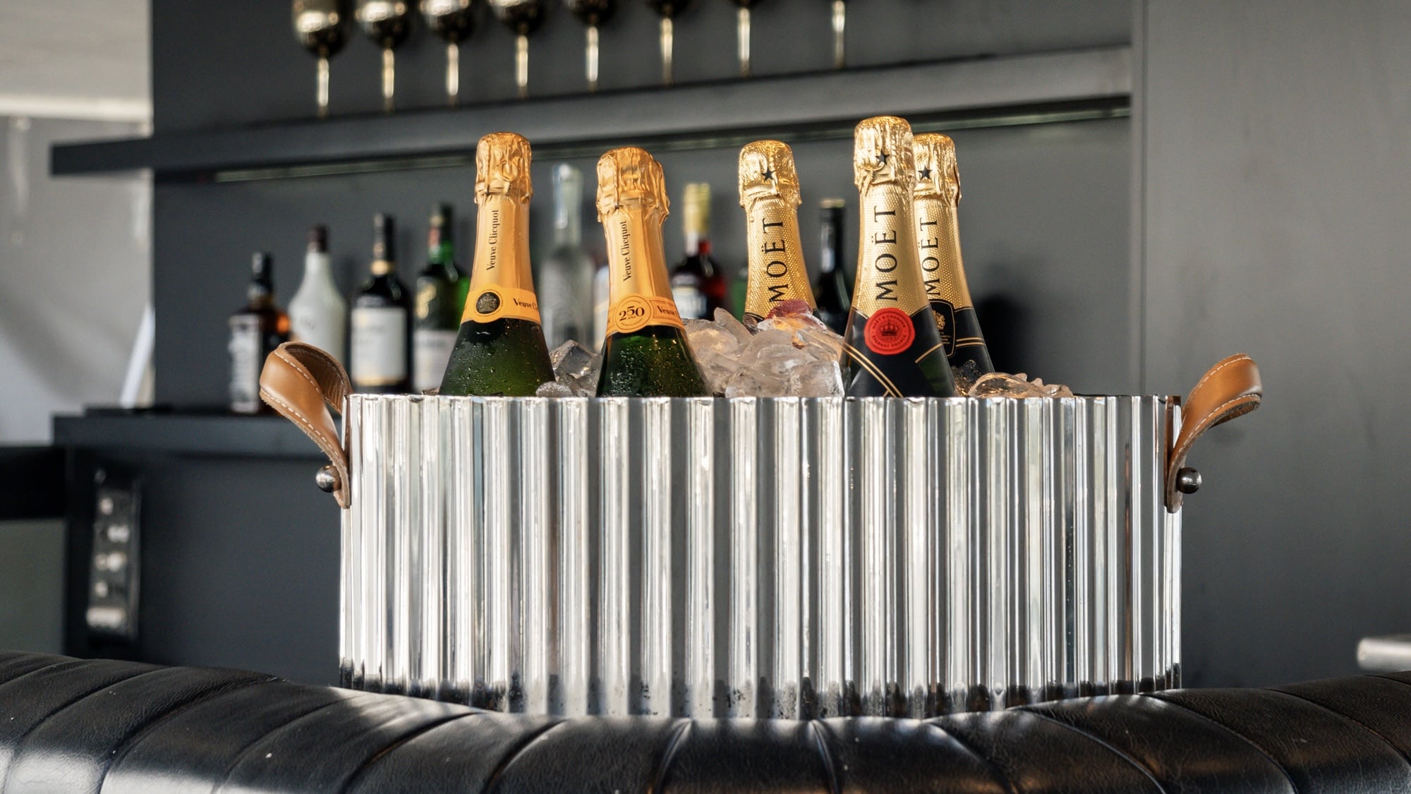 A metal ice bucket filled with bottles of Aussie Magic champagne sits on a leather surface, while liquor bottles and glasses shimmer on shelves in the background.