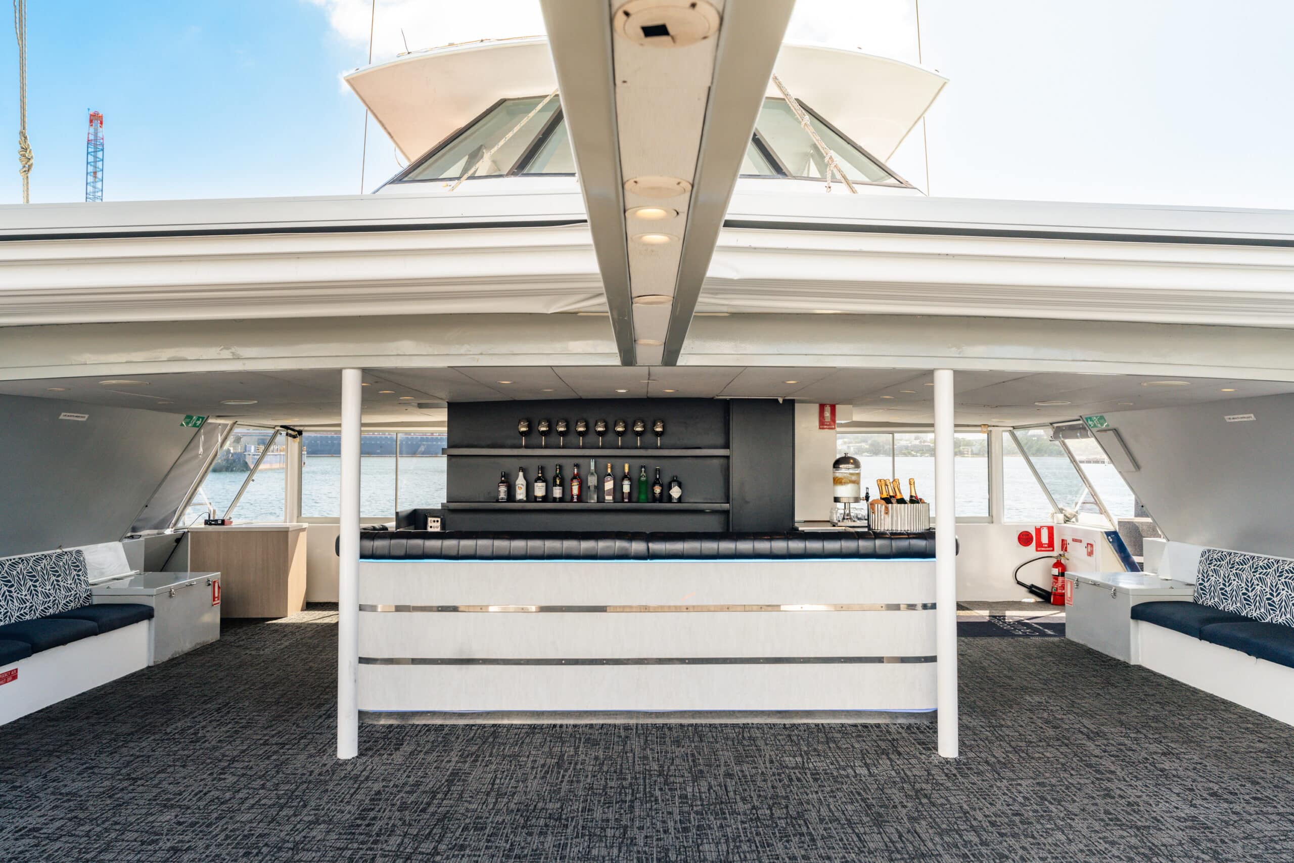 Aussie Magic’s modern yacht interior features a bar with bottle displays, cushioned side seating, large windows offering water views, and carpeted floors filled with natural light.