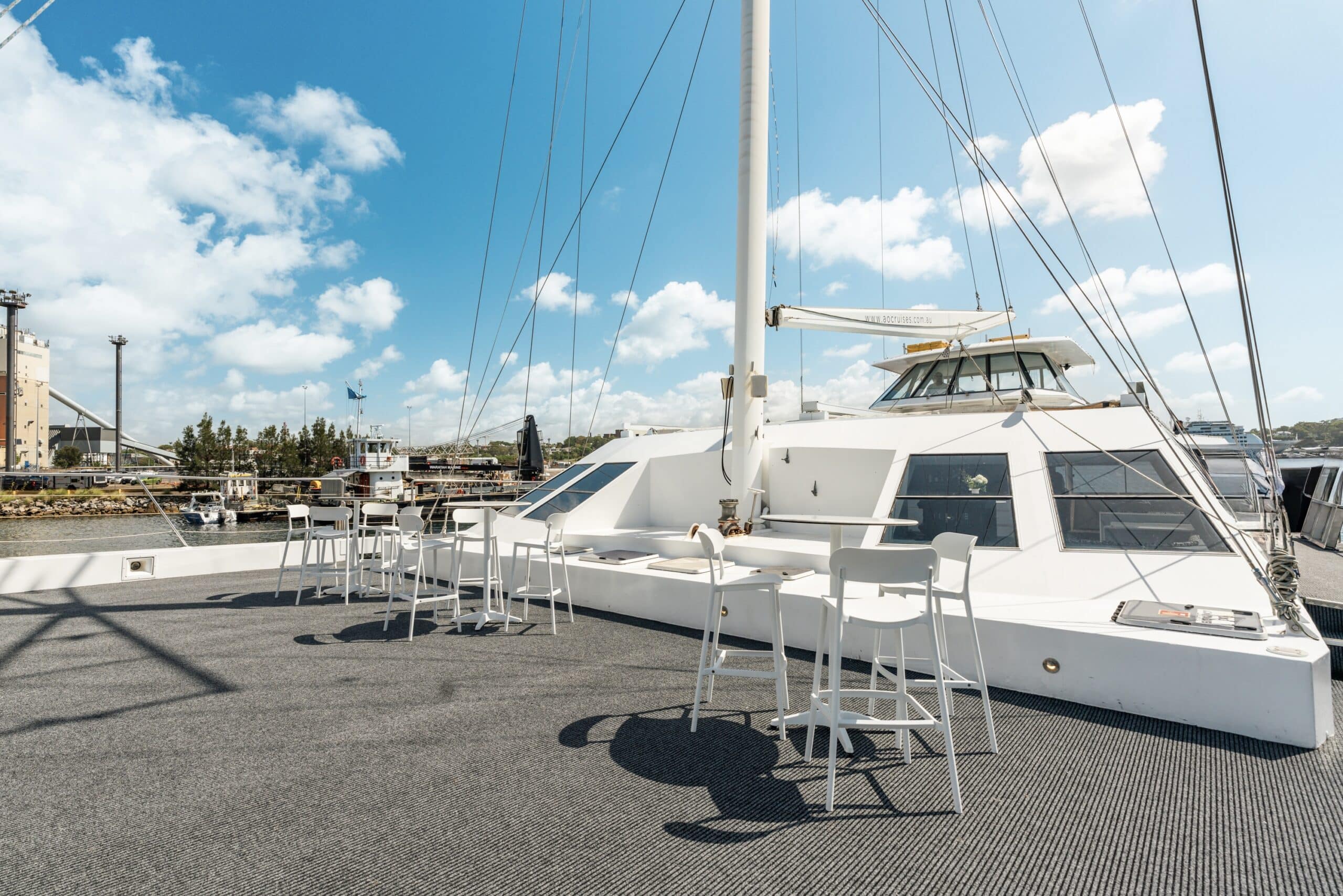 The Aussie Magic yacht features a sunny outdoor deck with high tables and chairs, offering harbor views of boats, buildings, and trees beneath a blue sky with scattered clouds.