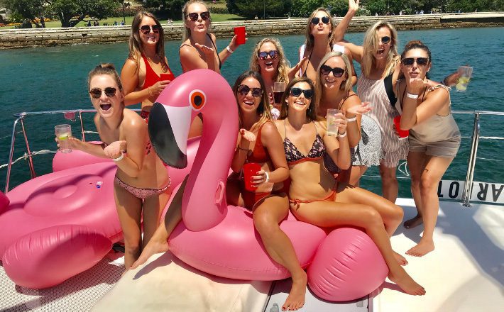 A group of nine women enjoy a sunny day on a boat, posing with drinks in hand next to a large inflatable pink flamingo. The women are smiling and wearing swimwear and sunglasses. The backdrop features water and a grassy shoreline with trees, courtesy of The Yacht Social Club Sydney Boat Hire.