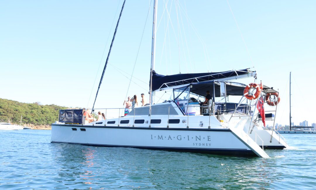 A white sailboat named "IMAGINE SYDNEY" is floating on a calm body of water. The boat, available for Sydney Harbour Boat Hire The Yacht Social Club, has multiple people on board and a forested shore in the background under a clear blue sky. Two red life rings are visible on the side of the boat.