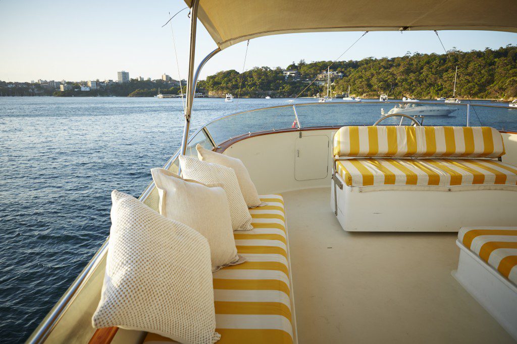 A boat's deck with cushioned seating featuring yellow and white striped upholstery, and white throw pillows. Docked near a scenic coastline with green hills and several boats anchored in the distance, it’s perfect for a Luxury Yacht Rentals Sydney experience. The sky is clear, and the water calm.