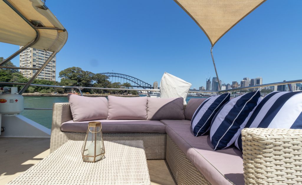 A cozy seating area on a luxury yacht with beige wicker furniture and striped pillows, overlooking a body of water. In the background, there is a city skyline with prominent skyscrapers and a bridge under a clear blue sky. A lantern decorates the coffee table. Experience this with The Yacht Social Club Sydney Boat Hire.