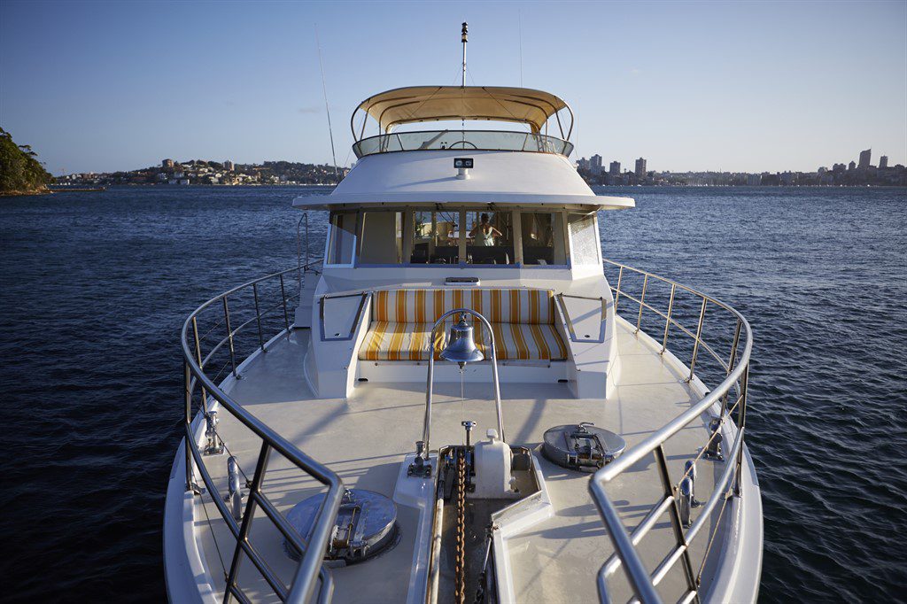 A yacht with a white deck and yellow-striped seating is anchored in calm waters with a cityscape in the background. The view from the front of the yacht showcases metal railings and various deck fixtures. The sky is clear and blue, perfect for Boat Parties Sydney The Yacht Social Club.