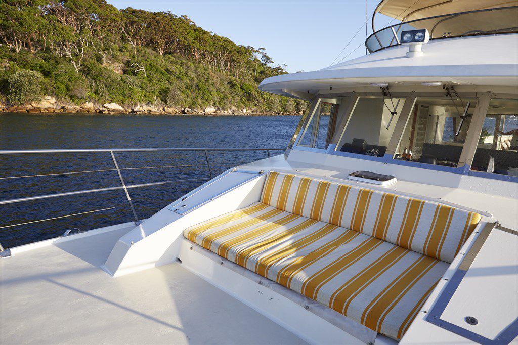 A comfortable seating area with yellow and white striped cushions is on the deck of The Yacht Social Club boat. The vessel is situated on calm water near a densely forested shoreline with clear skies in the background, making it ideal for a Sydney Harbour Boat Hire experience.