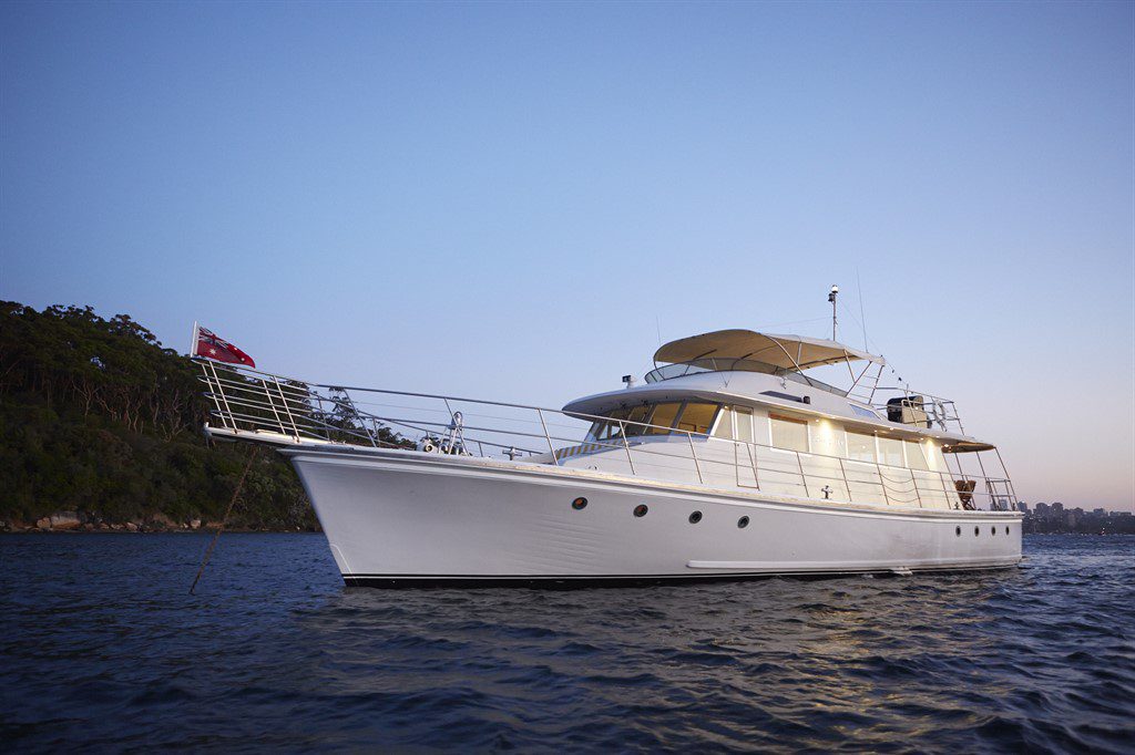 A luxurious white yacht with multiple decks is moored on calm waters during twilight. The vessel, perfect for Boat Parties Sydney by The Yacht Social Club, features railings, a canopy on the upper deck, and a red flag at the stern. Trees and a rocky shoreline are visible in the background under a clear sky.