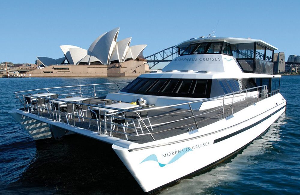 A luxurious white yacht named "Morpheus Cruises" floats on clear blue waters, a perfect example of The Yacht Social Club Sydney Boat Hire. The Sydney Opera House, with its iconic sail-like design, stands prominently in the background under a cloudless sky.