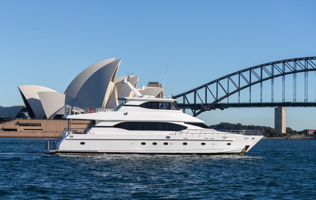 A white yacht cruises through the water with the Sydney Opera House and Sydney Harbour Bridge in the background on a clear, sunny day, exemplifying The Yacht Social Club's premium Sydney boat hire experience.