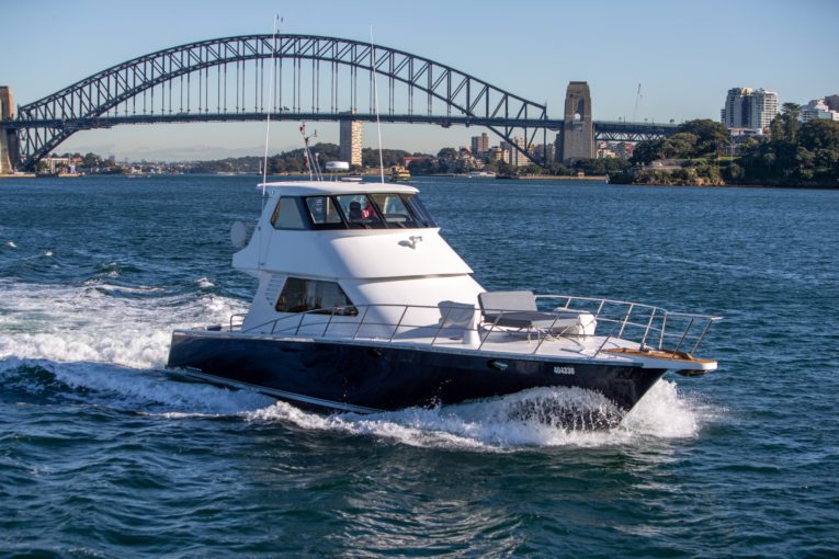 A white motor yacht from The Yacht Social Club Sydney Boat Hire cruises on clear blue waters with the iconic Sydney Harbour Bridge in the background under a clear sky. The boat leaves a wake as it moves forward, with urban buildings and greenery visible on the shore.
