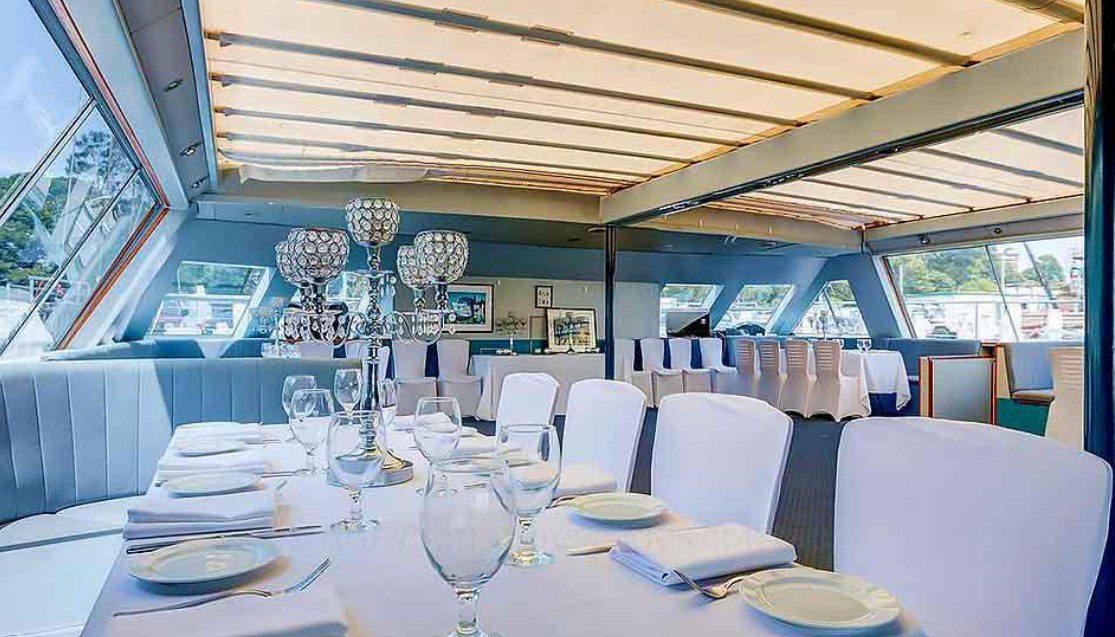 Aussie Magic features an elegant dining area with white tablecloths, glassware, and crystal candelabras. Large windows offer natural light and scenic views of the surrounding water and greenery.