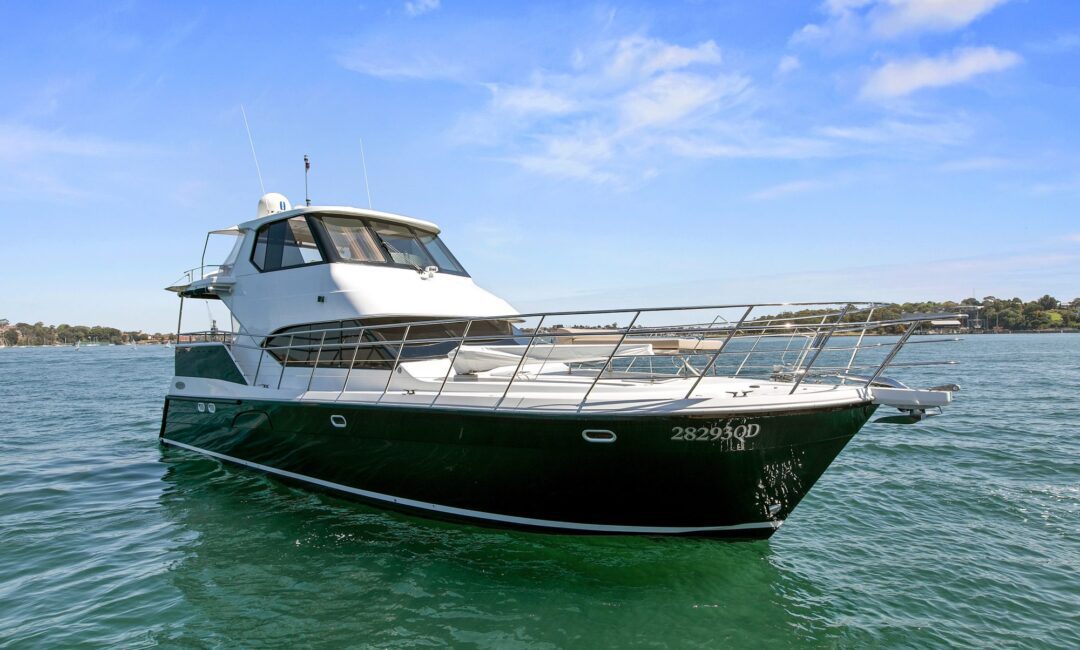 A large luxury yacht with a white upper deck and a dark green hull is anchored on calm waters. The boat features multiple antennas on the roof and railings along the deck. In the background, there is a shoreline with trees and buildings under a clear blue sky—a perfect scene for Luxury Yacht Rentals Sydney.