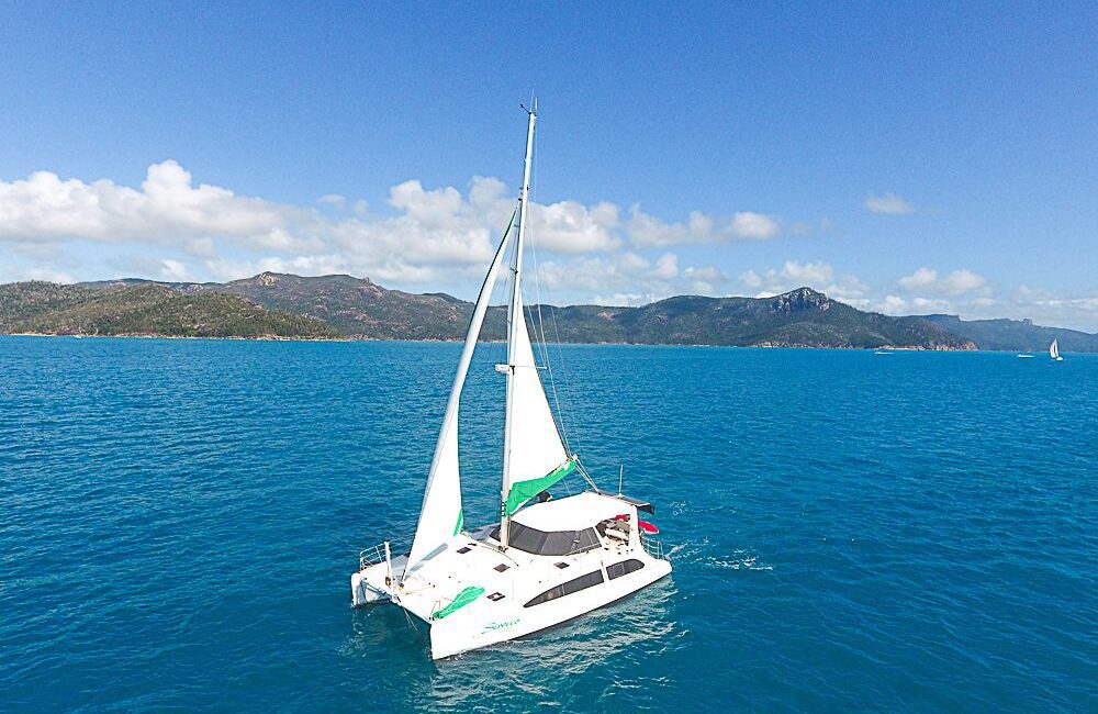 A white catamaran with green accents sails in clear blue waters on a sunny day. Hills covered in greenery are visible in the distance under a partly cloudy sky. Another sailboat can be seen far off in the background, adding to the picturesque setting perfect for The Yacht Social Club Event Boat Charters.