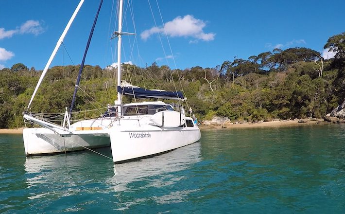 A white catamaran named "Woorobinda" is anchored in clear turquoise waters near a forested shoreline with a sandy beach. The sky is clear with a few scattered clouds. Experience this serene escape with Boat Rental and Parties Sydney The Yacht Social Club.