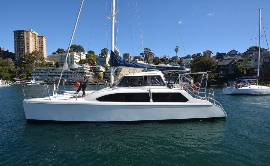 A white sailboat with a blue sail is docked on a calm body of water, surrounded by tall residential structures and waterfront houses. A few people are on the boat under a clear sky. Enjoy this serene scene with The Yacht Social Club Sydney Boat Hire for unforgettable boating experiences.