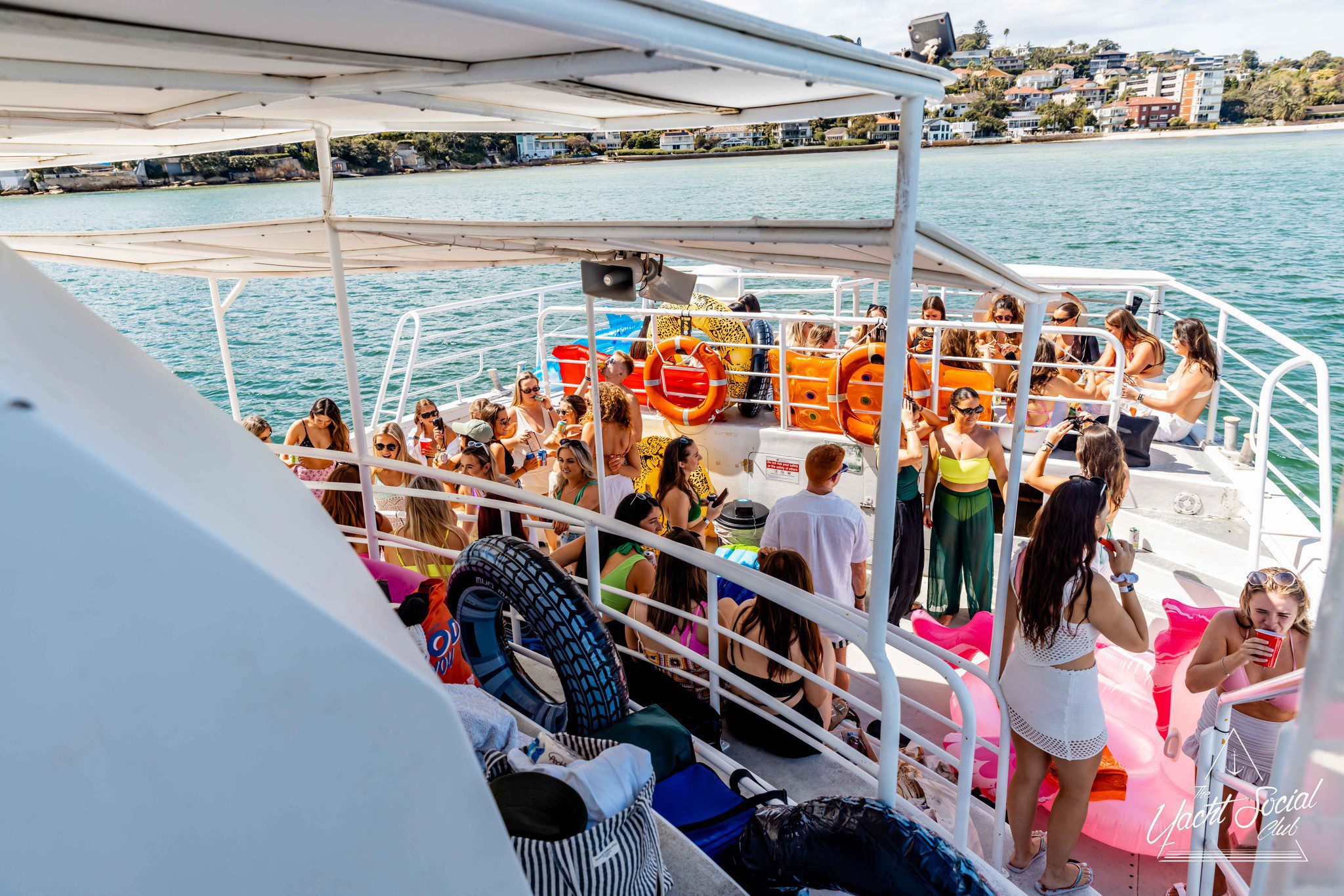 A group of people enjoy a sunny day on a boat, with various inflatable toys around them, against a backdrop of a calm sea and distant coastline.