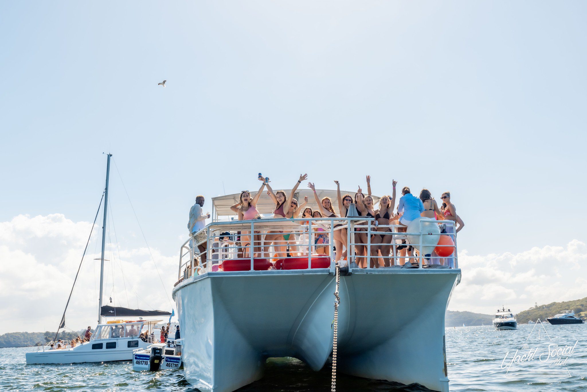 A group of people are standing on the deck of a large boat, waving and posing for a photo. Other boats are visible in the background on a sunny day.