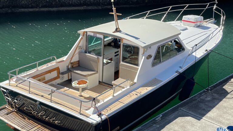 A modern boat is docked at a pier. It features a white cabin with windows and seating, including a small table, in the deck area. The boat's name, "El Soliste," is visible on the stern. The water around the boat is clear green, and the concrete pier enhances the setting. Ideal for Boat Parties Sydney The Yacht Social Club.