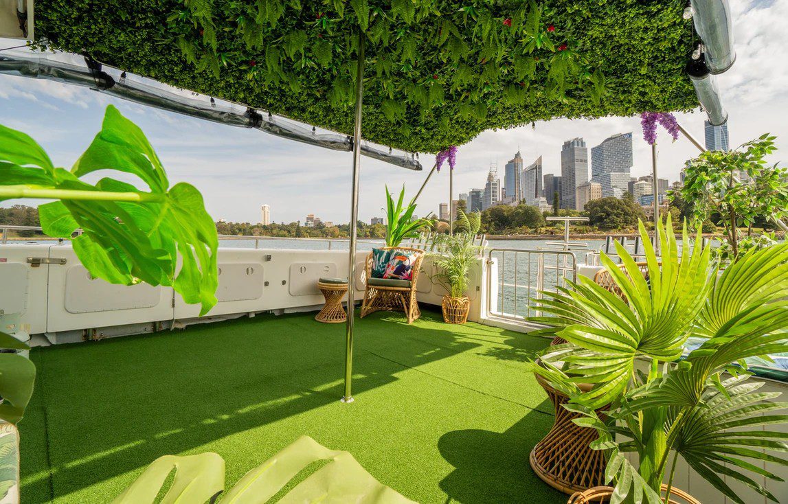 A rooftop deck adorned with lush greenery, including potted plants and a green, leaf-covered canopy. The space has wicker furniture and a backdrop of a city skyline with high-rise buildings. Blue skies and bright sunlight enhance the serene atmosphere, making it perfect for The Yacht Social Club Event Boat Charters.