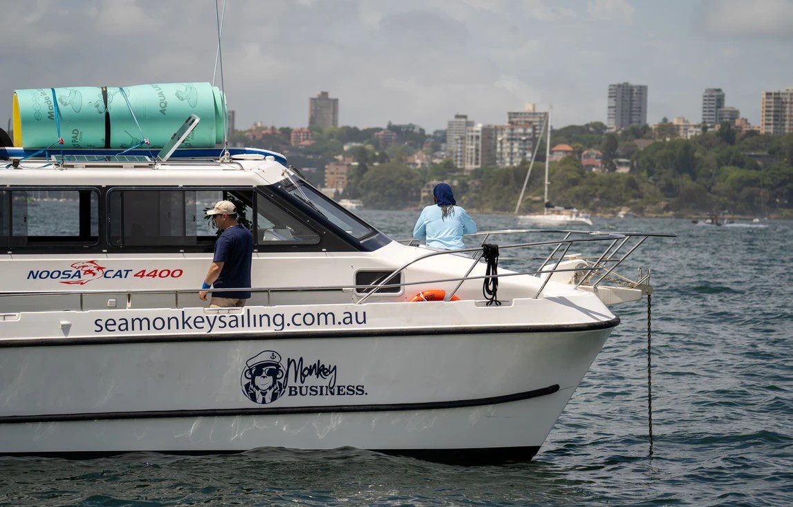 A white boat labeled "seamonkeysailing.com.au" and "Monkey Business" is on the water. Two people are on board: one is standing on the side and the other is near the cabin. The coastline with buildings and trees is visible in the background, perfect for a day of Sydney Harbour Boat Hire The Yacht Social Club.