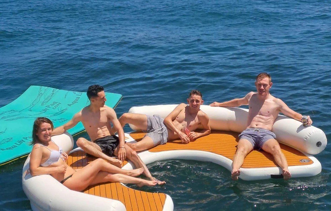 Four people are relaxing on a floating white and brown inflatable lounge in clear blue water. A woman and three men, all dressed in swimwear, are smiling and enjoying a sunny day. A green floating mat is visible in the background, perfectly capturing the essence of The Yacht Social Club Sydney Boat Hire.