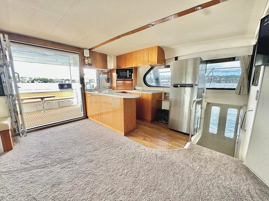 Interior of a yacht featuring a modern kitchen with wooden cabinets, stainless steel appliances, and a microwave. A sliding glass door opens to an outdoor seating area. The carpeted floor transitions to wooden flooring near the kitchen. Large windows offer sea views.