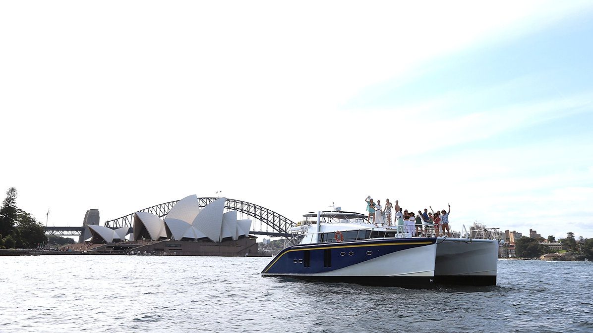 A modern white and blue catamaran with people on board sails near the iconic Sydney Opera House and Harbour Bridge on a sunny day. The water is calm, and the passengers appear to be enjoying the scenic views.