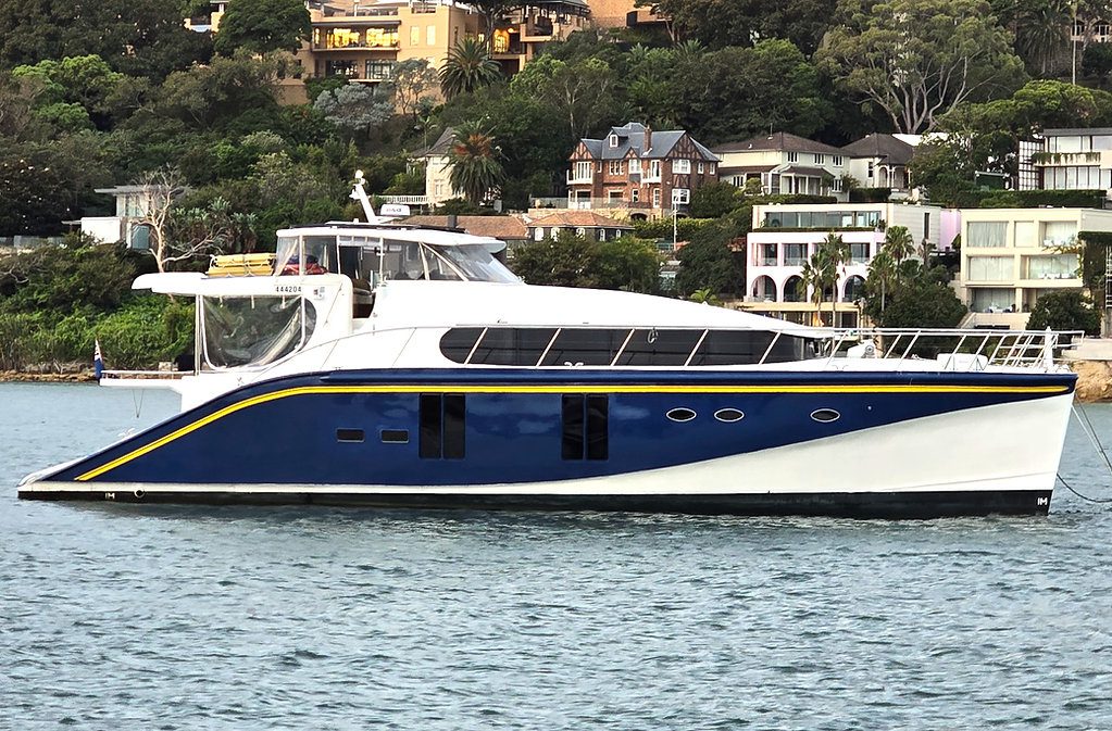 A sleek, blue and white motor yacht floats on water with a backdrop of upscale houses and greenery on a hillside. The yacht has a modern design with large windows and a spacious deck area.
