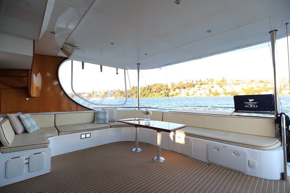 The image shows the interior of a yacht's deck, featuring a spacious seating area with beige cushions and two small pillows. A central table has a single flower vase, and the large windows reveal a scenic view of the water and shoreline outside.