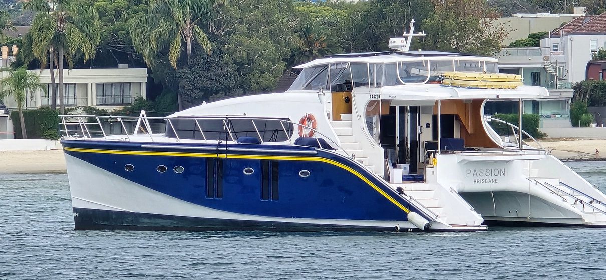 A sleek blue and white luxury yacht named "Passion" is anchored in a calm waterway near a residential area with modern houses and lush trees in the background. The yacht features an upper deck and several portholes on the side.