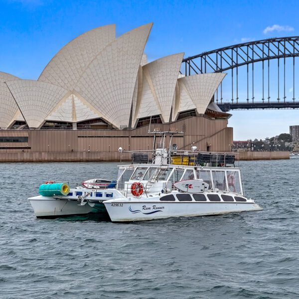 Rum Runner floats on the water in front of the iconic Sydney Opera House and Sydney Harbour Bridge. The sky is clear with a few clouds, and the urban skyline is visible in the background. The boat has several colourful objects on its deck.