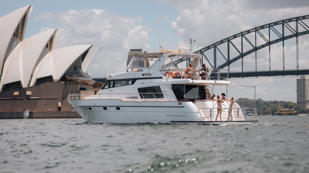 A white yacht cruises near the Sydney Opera House and Sydney Harbour Bridge. Several people are seen relaxing on the deck of the boat, enjoying the sunny weather and scenic views. The water is calm, and the sky is partially cloudy.