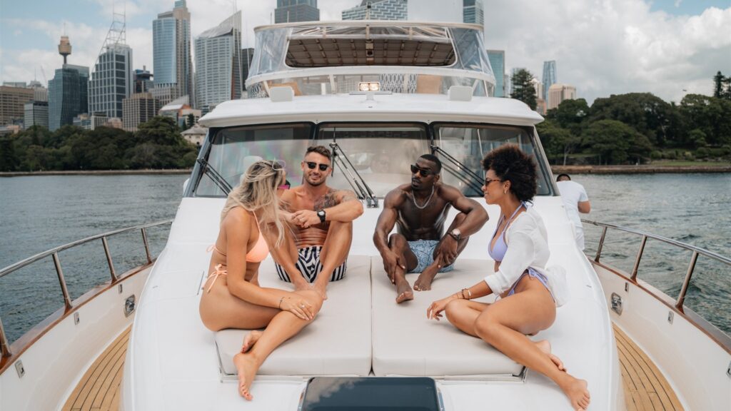 A group of four people relaxing on a yacht's deck with a city skyline in the background. Two women in bikinis and two men in swim trunks are sitting and chatting under a partly cloudy sky. The cityscape features tall buildings and lush greenery.