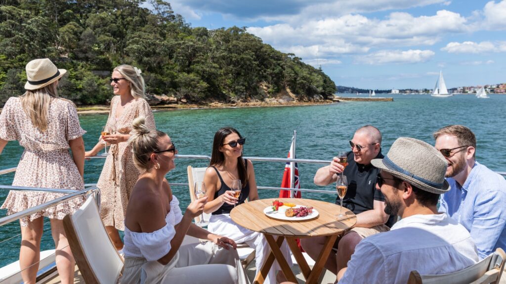 A group of six people enjoys a sunny day on a boat, smiling and mingling. Some are seated at a table with drinks and appetizers, while others stand by the railing. The backdrop includes a serene body of water, lush greenery, and a distant sailboat under a partly cloudy sky.