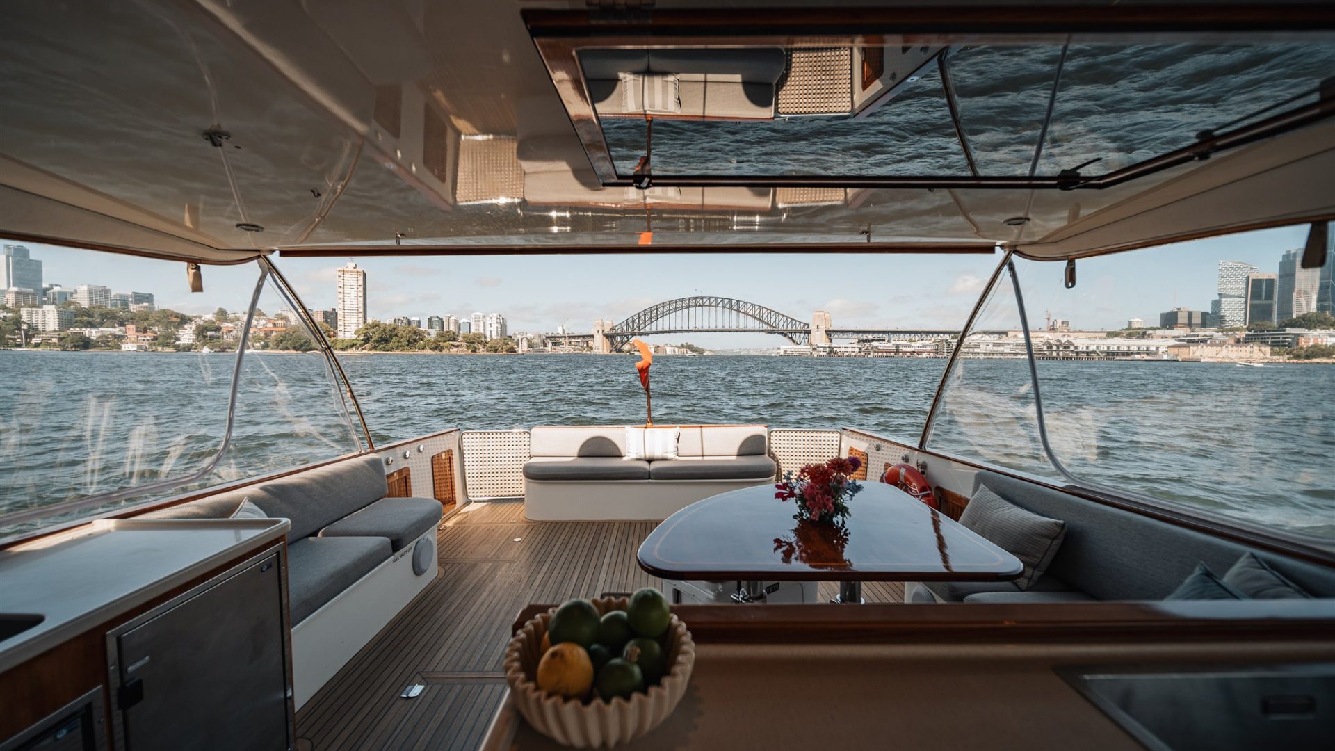 Interior view from inside a yacht, showcasing a luxurious seating area and a dining table with a flower centerpiece. Through the open rear, a city skyline and a prominent bridge are visible across a body of water. The scene is well-lit with natural daylight.