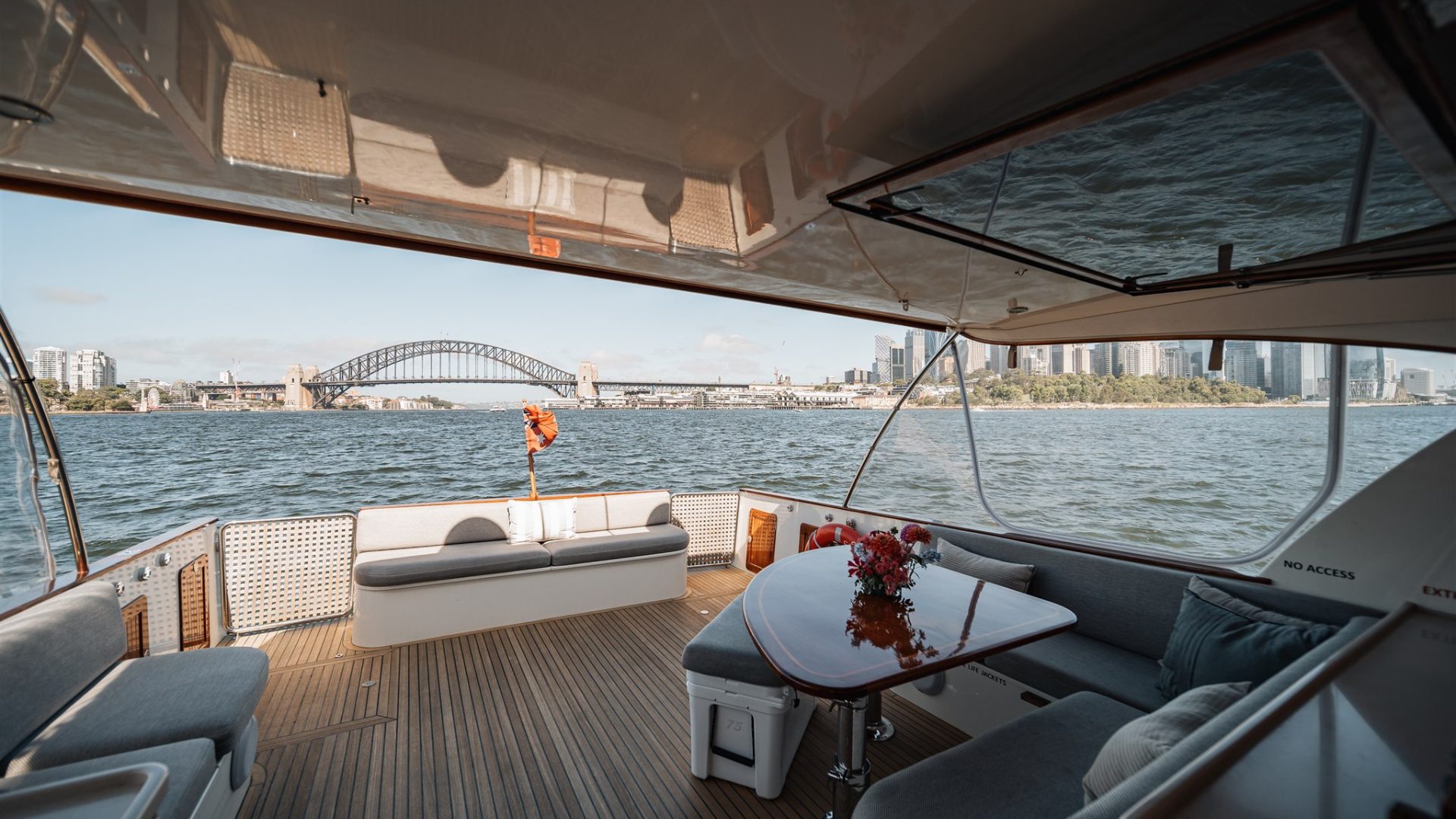 A view from inside a luxurious boat looking out towards a large body of water with the Sydney Harbour Bridge in the background. The boat's interior includes seating, a table with flowers, and a partially open deck. The skyline of Sydney is visible on the right.