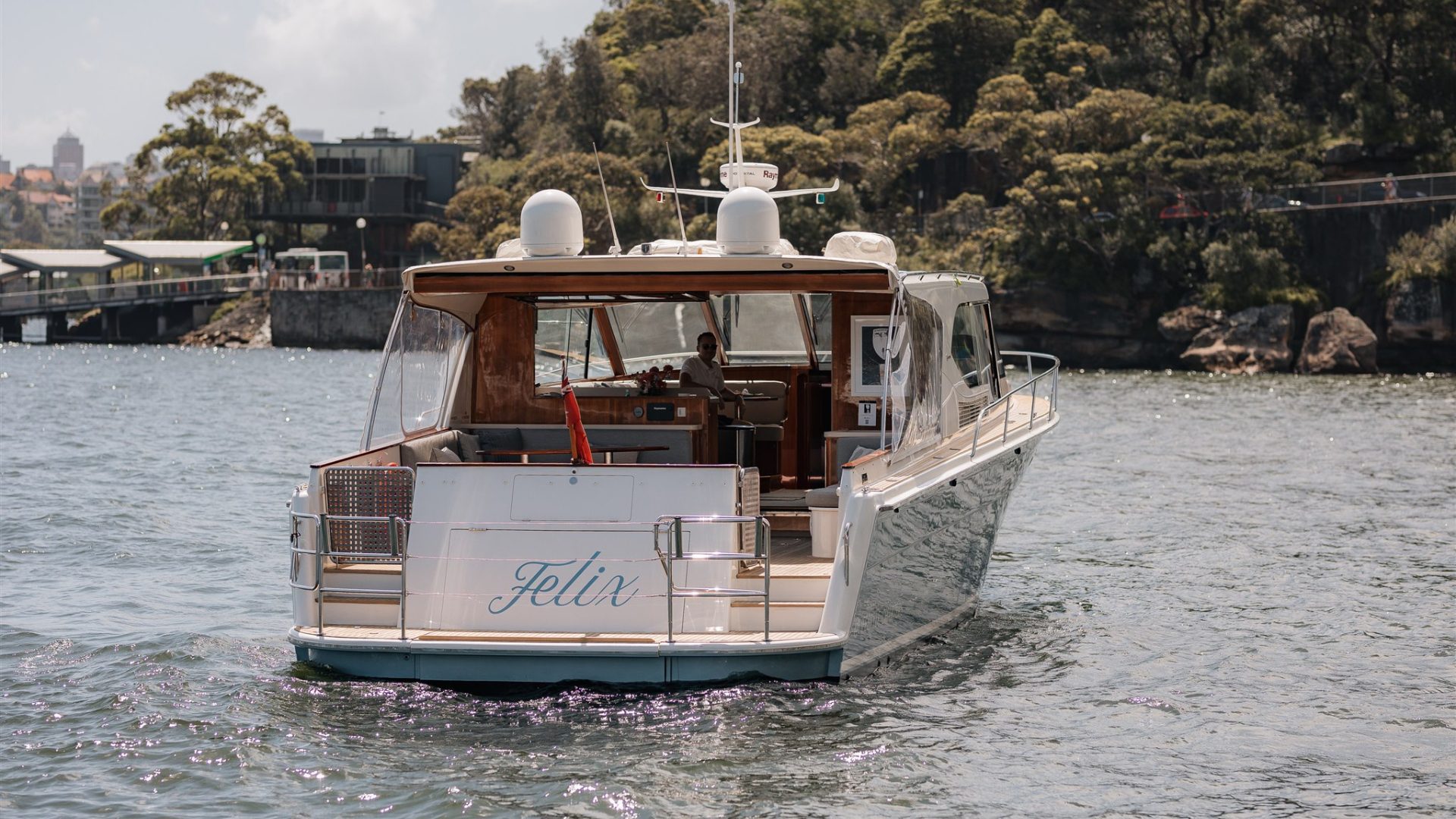 A motorboat named "Felix" is anchored on a calm body of water, with a lush, green, forested shoreline in the background. The boat's rear is visible, showcasing its deck and interior seating area. Additionally, a building is visible on the left side of the shoreline.