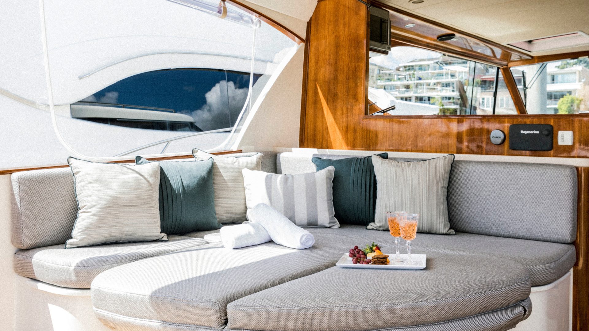 Comfortable seating area on a yacht with gray cushioned benches, green and white pillows, and a small tray with two glasses of rosé wine, grapes, and cheese on the table. Natural light pours in, highlighting the wooden interiors and marina visible outside.