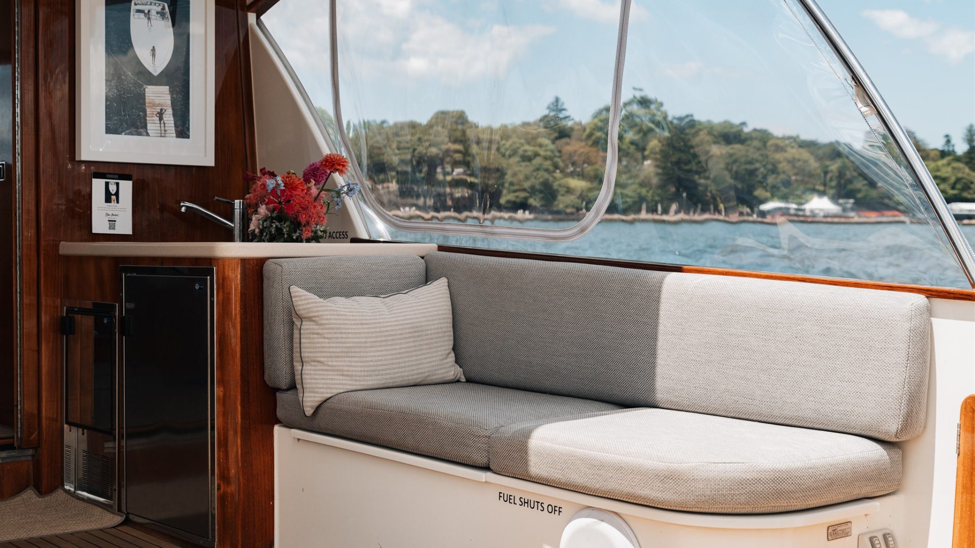 Interior of a boat featuring a cozy seating area with a light grey cushioned bench, a decorative pillow, a small counter with flowers in a vase, and a framed picture on the wall. Large windows provide a picturesque view of a lake and trees outside.