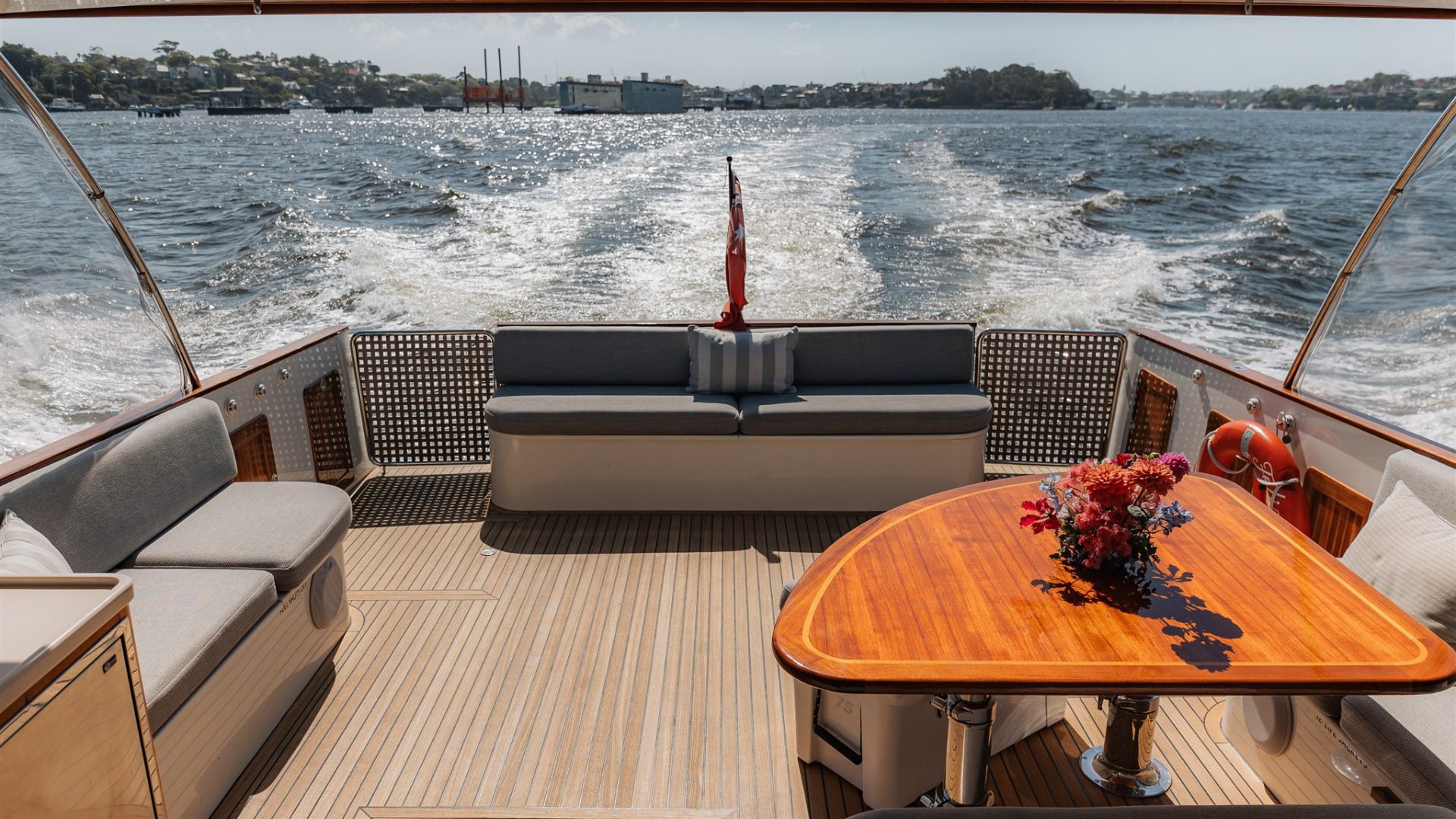 The image shows the rear deck of a boat with wooden flooring and cushioned seating areas along the sides. A polished wooden table with a bouquet of flowers is in the center. The boat is moving on a large body of water, and buildings and greenery are visible in the background.