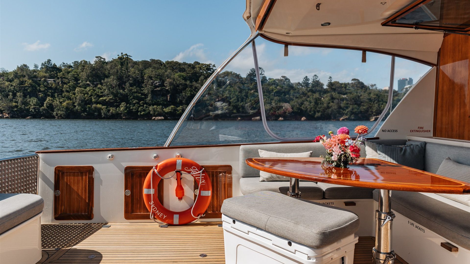 A view of a boat's seating area with a wooden table topped with a bouquet of flowers, cushioned benches, and a life preserver hanging on the railing. The boat is on a body of water with a forested shoreline in the background under a clear sky.