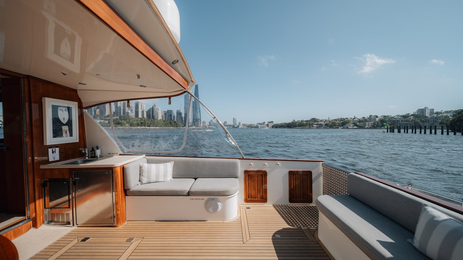 View from the deck of a yacht with a wooden floor and cushioned seating. The city skyline and trees are visible in the distance under a clear, sunny sky. The setup includes a small counter and framed artwork on the interior wall, offering a luxurious, serene setting.