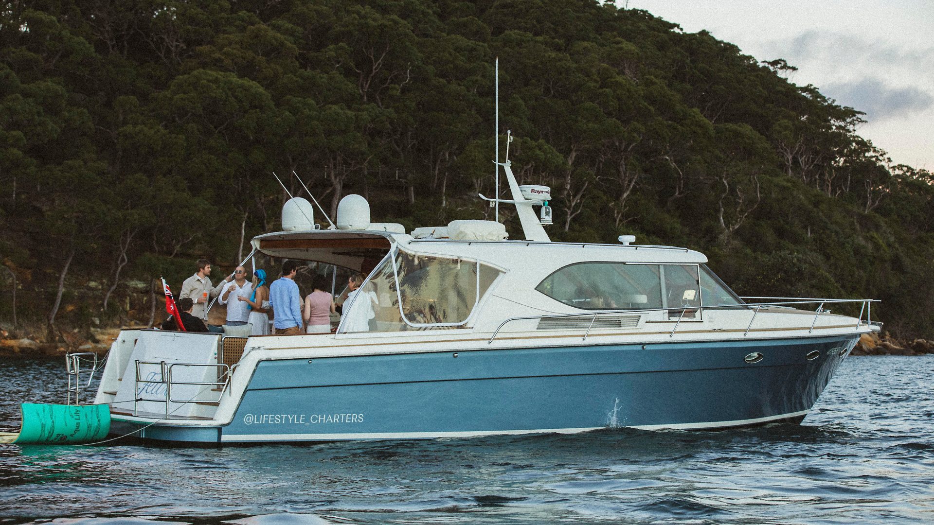 A group of people are gathered on the deck of a blue and white yacht named "LIFESTYLE CHARTERS" anchored near a forested shoreline. The yacht has a spacious deck with comfortable seating and a few steps leading to the water.