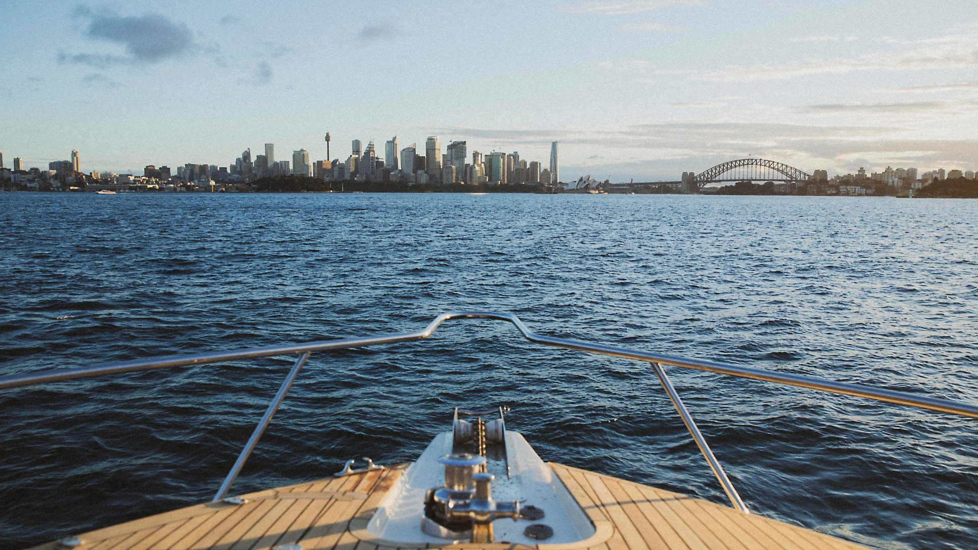 The view from a boat's deck shows a sprawling city skyline across a body of water, with a prominent bridge and a tower visible against the sky at dusk. The scene reflects a calm and serene aquatic journey toward an urban landscape.