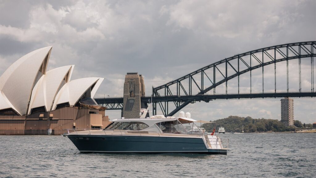 A luxury boat on Sydney Harbour is cruising near the iconic Sydney Opera House and the Sydney Harbour Bridge on an overcast day. The boat is sleek and modern, with a few people seen on board enjoying the view.