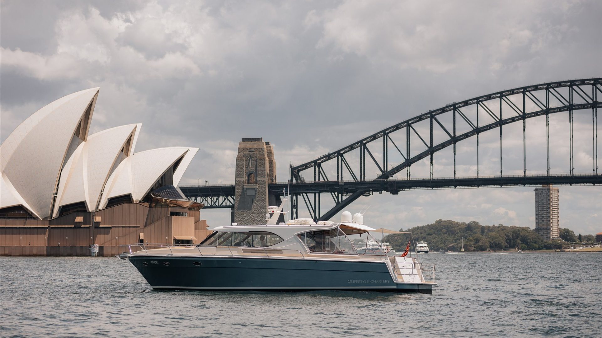 A luxury boat on Sydney Harbour is cruising near the iconic Sydney Opera House and the Sydney Harbour Bridge on an overcast day. The boat is sleek and modern, with a few people seen on board enjoying the view.