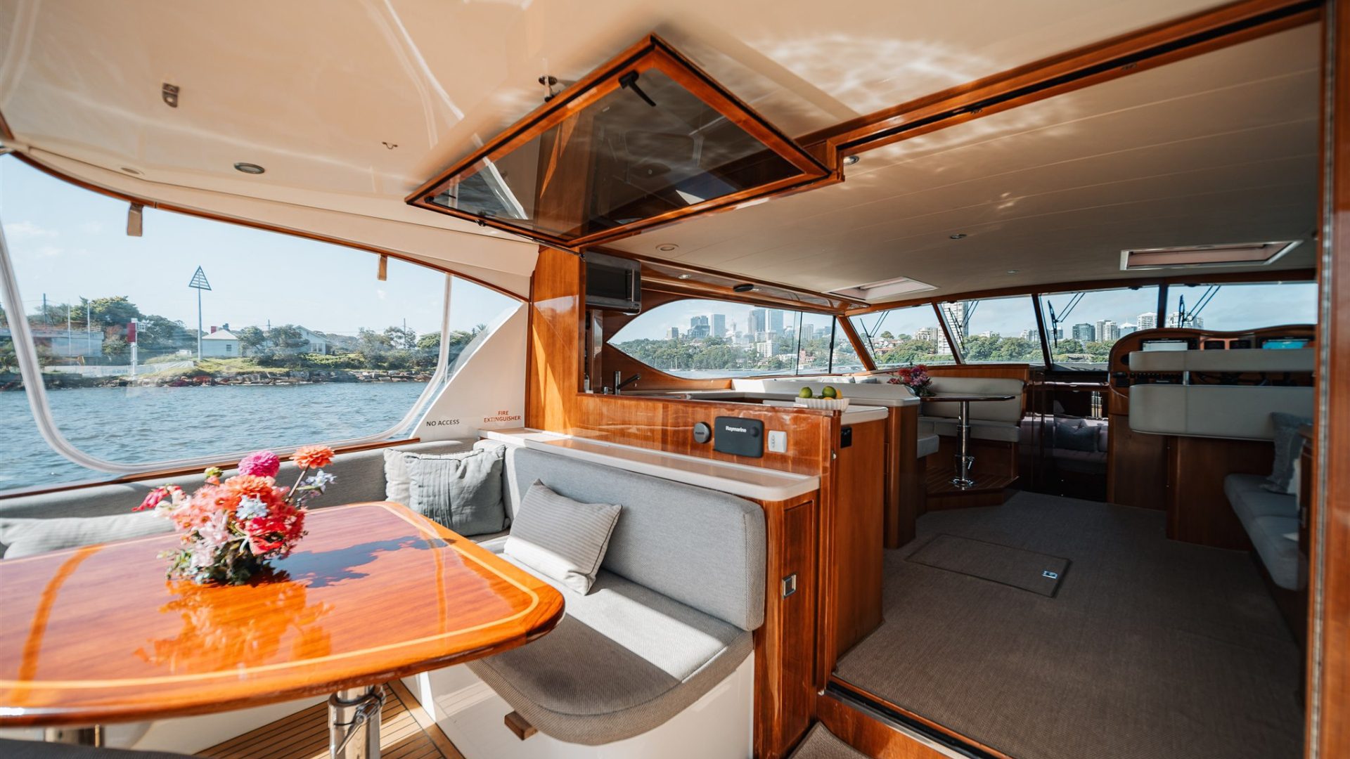 The image shows the interior of a luxury yacht with polished wooden furnishings. There is a table with a vase of flowers, cushioned seating, a kitchenette, and large windows offering a view of the water and cityscape. The space is sunlit and inviting.
