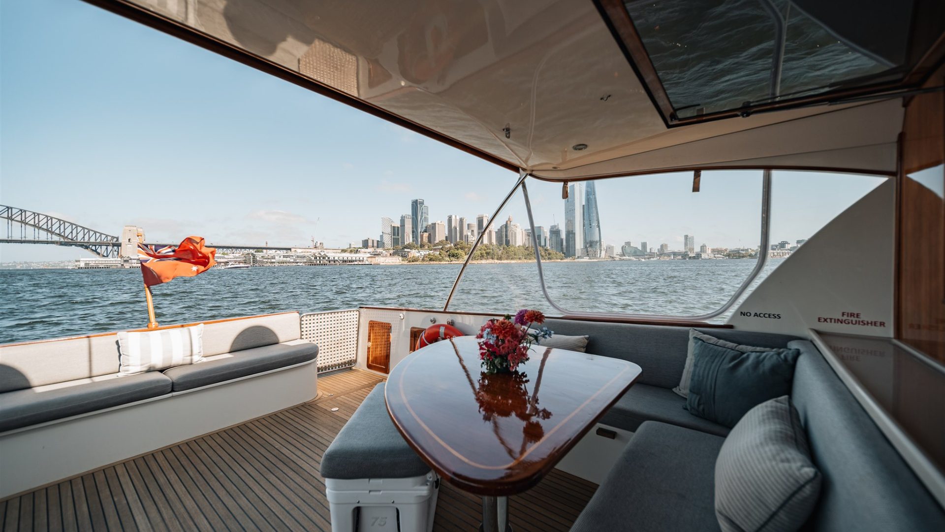 View from inside a boat showing a comfortable seating area with a wooden table topped with flowers. The boat is on water, and the background features a vibrant city skyline, including a bridge and skyscrapers under a clear blue sky.