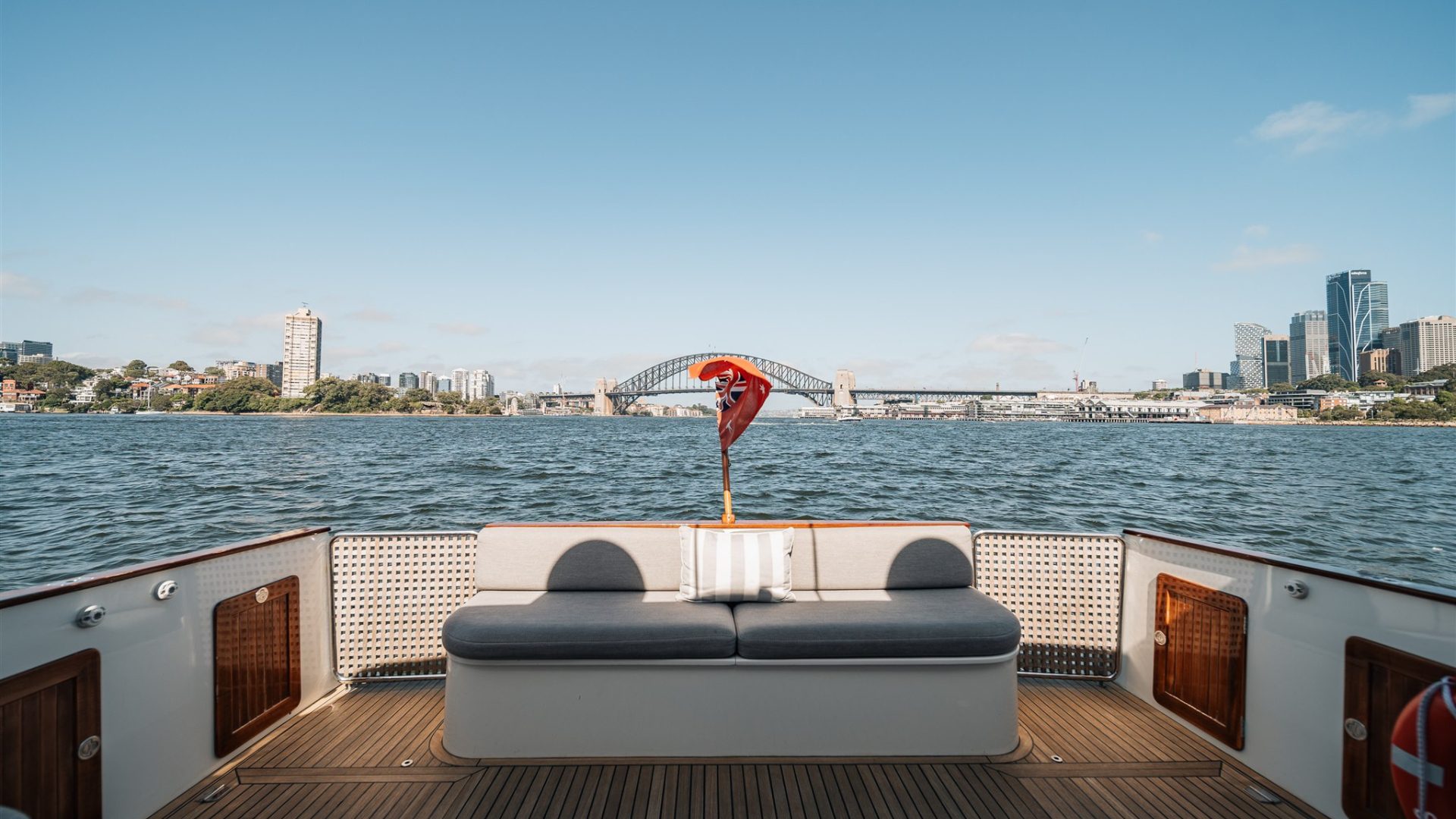 View from the deck of a boat looking out at the water with a city skyline in the background. The deck has a cushioned bench seating, a red and white life jacket hanging in the center, and a bridge visible in the distance. The sky is clear and blue.