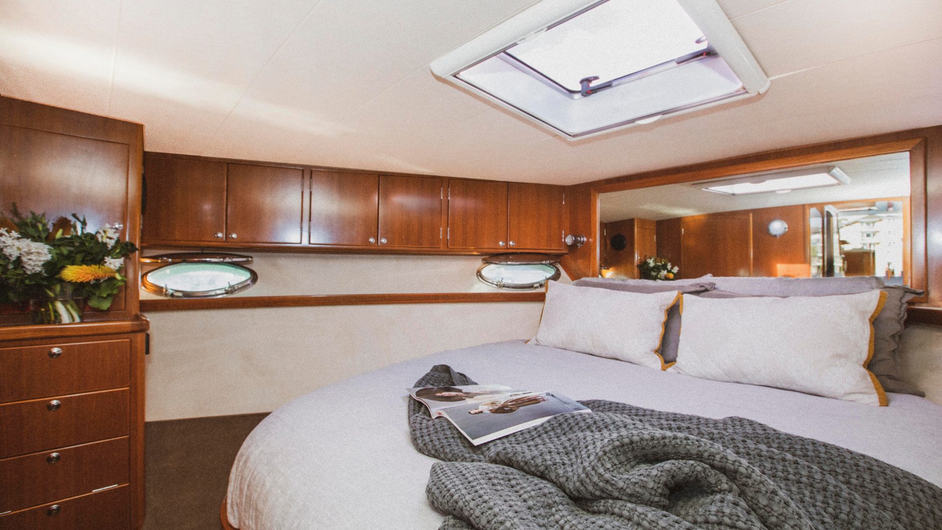 A cozy bedroom in a boat with a large bed covered in light gray linens and gray pillows. A magazine and a gray blanket are on the bed. The room features wooden cabinets, two round porthole windows, and a skylight. There's fresh flower arrangements on the left side.