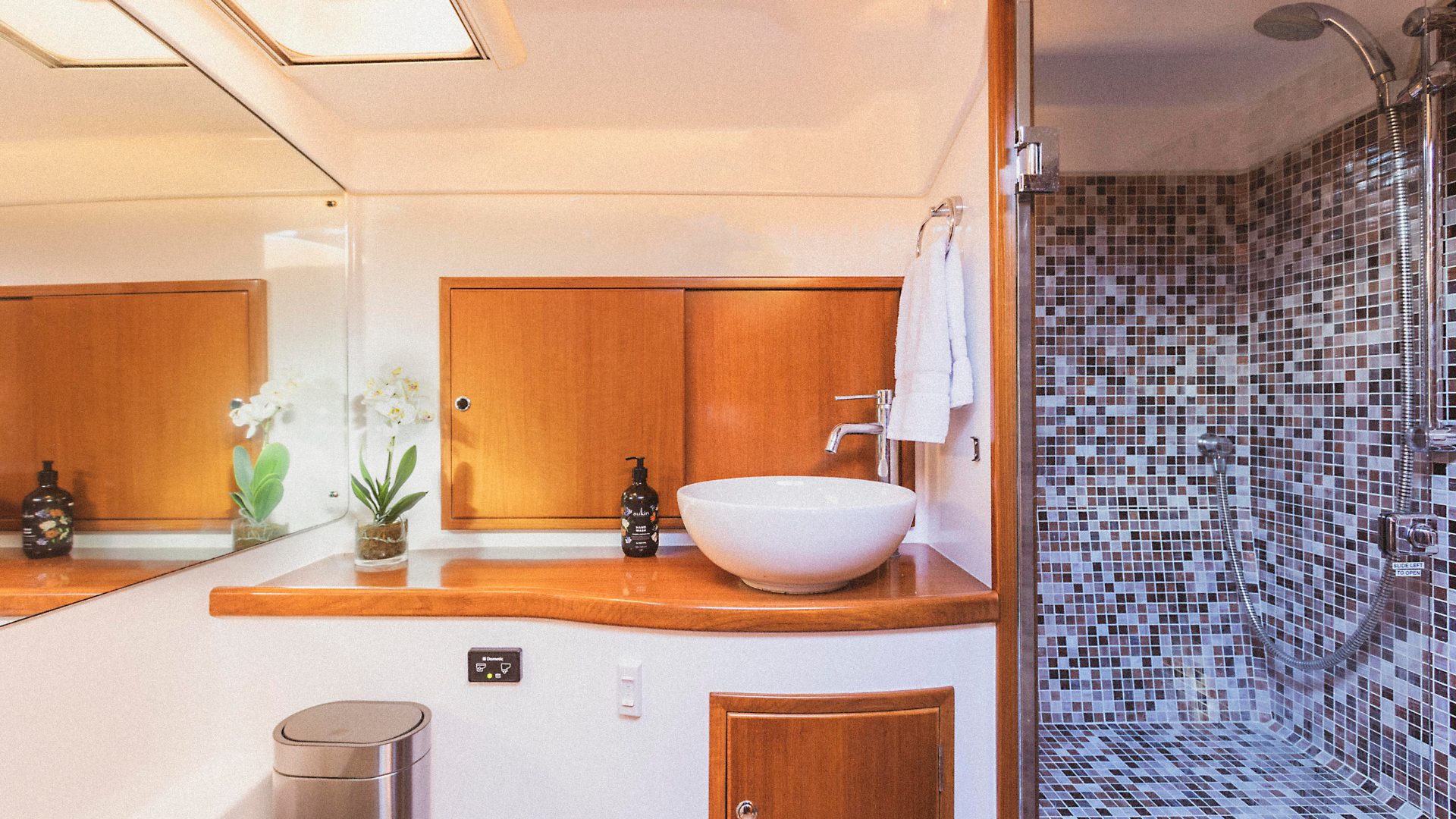 The image shows a modern bathroom with a wooden countertop, a white vessel sink, a mirrored wall, and a flower pot with white orchids. The shower area has a glass door and mosaic tile walls in shades of gray and white. Towels hang on a wall-mounted rack.